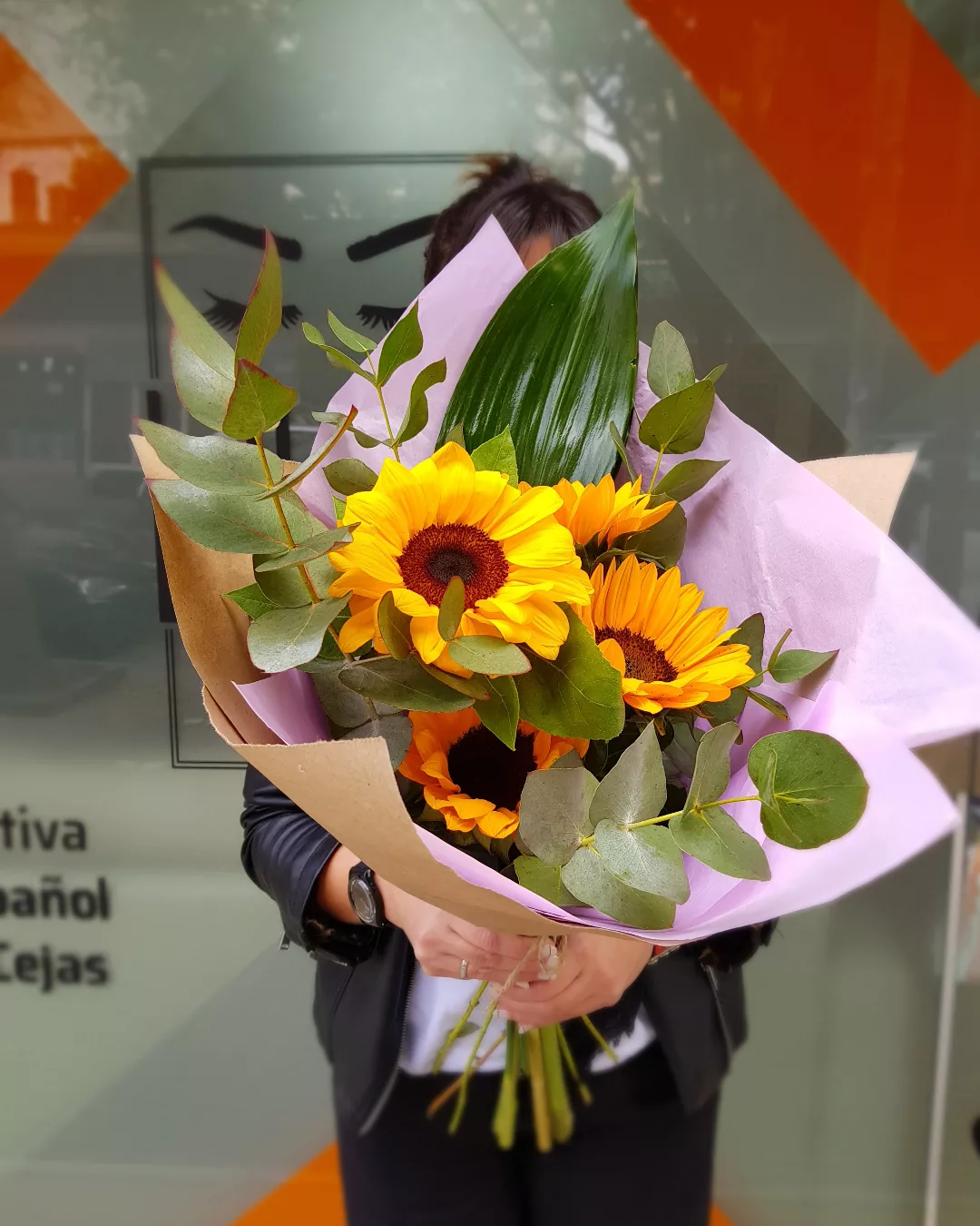 Nuestra vidriera, con el logo de los ojos y las franjas naranjas, y una clienta que se lleva un ramo de girasoles